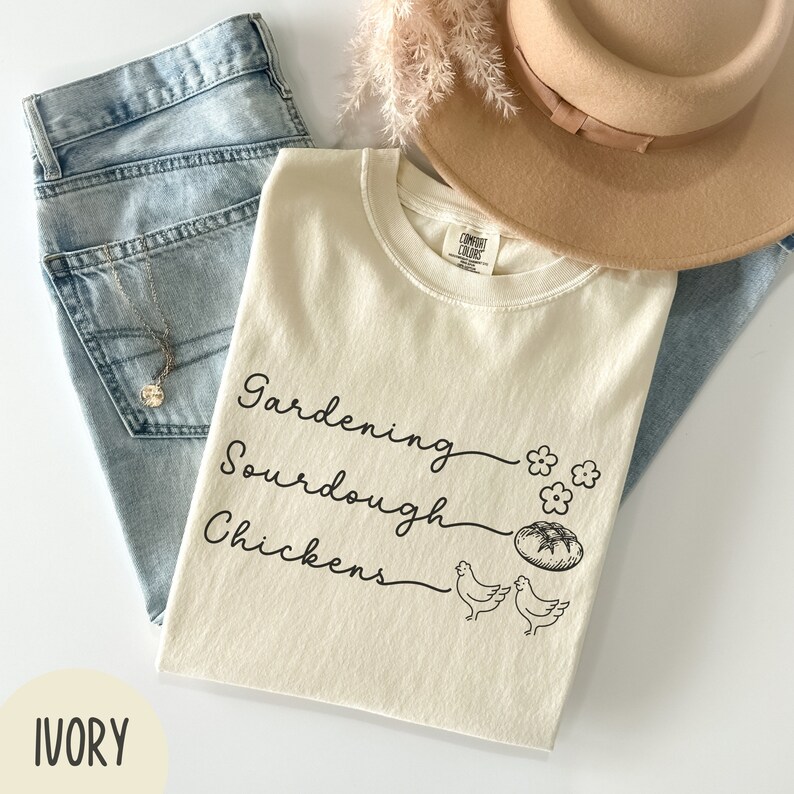 May include: Ivory t-shirt with the text "Gardening, Sourdough, Chickens" in black script. The shirt features a small illustration of a loaf of bread and two chickens.