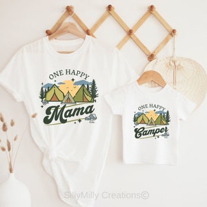 Puede incluir: Dos camisetas blancas con tem&aacute;tica de camping. La camiseta m&aacute;s grande dice "One Happy Mama" con un gr&aacute;fico de una puesta de sol, monta&ntilde;as y tiendas de campa&ntilde;a. La camiseta m&aacute;s peque&ntilde;a dice "One Happy Camper" con el mismo gr&aacute;fico.