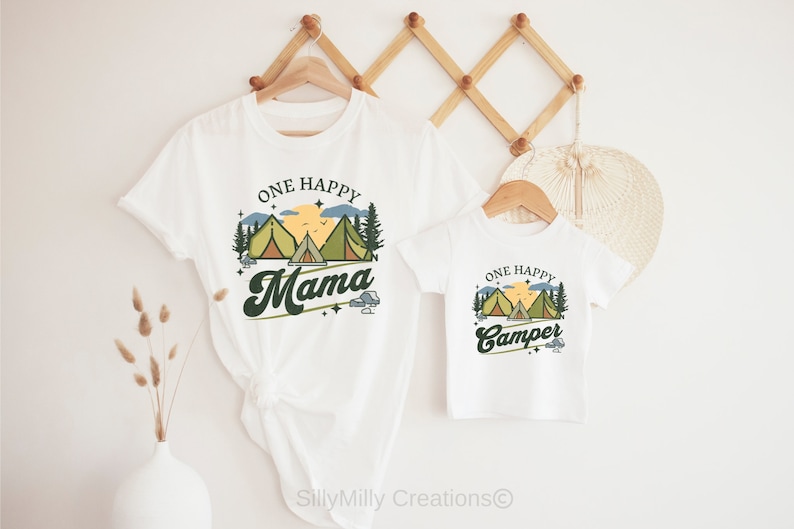 Puede incluir: Dos camisetas blancas con tem&aacute;tica de camping. La camiseta m&aacute;s grande dice "One Happy Mama" con un gr&aacute;fico de una puesta de sol, monta&ntilde;as y tiendas de campa&ntilde;a. La camiseta m&aacute;s peque&ntilde;a dice "One Happy Camper" con el mismo gr&aacute;fico.