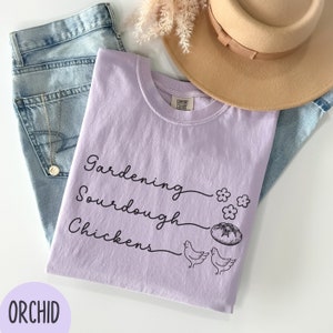 May include: A lavender t-shirt with the text "Gardening, Sourdough, Chickens" in black script. The shirt has a small graphic of a loaf of bread, flowers, and two chickens.