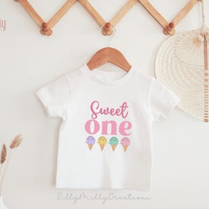 May include: White short-sleeved t-shirt with the text "Sweet one" in pink lettering and a row of five colorful ice cream cones below.