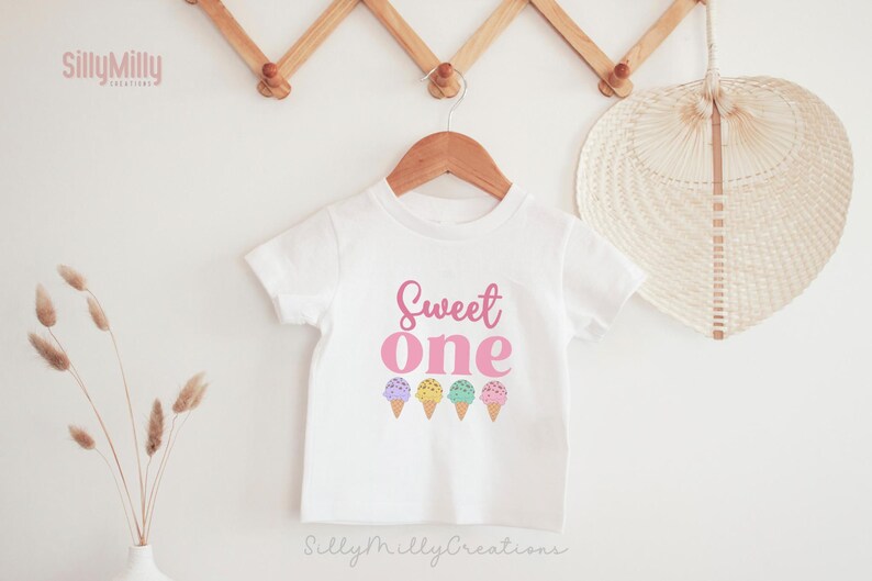 May include: White short-sleeved t-shirt with the text "Sweet one" in pink lettering and a row of five colorful ice cream cones below.