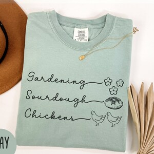 May include: A light green t-shirt with the text "Gardening, Sourdough, Chickens" in black script. The shirt has a small flower graphic and a bread graphic.