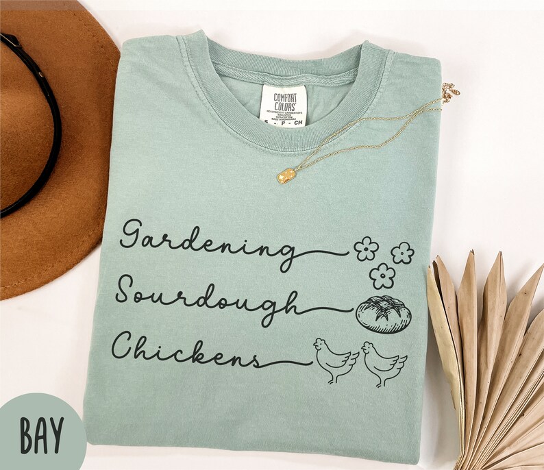 May include: A light green t-shirt with the text "Gardening, Sourdough, Chickens" in black script. The shirt has a small flower graphic and a bread graphic.