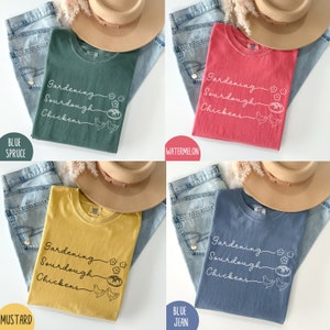 May include: Four different coloured t-shirts with the text "Gardening Sourdough Chickens" and a small illustration of a loaf of bread and two chickens. The shirts are displayed on a denim background with a hat.