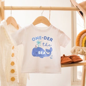 May include: White baby onesie with a blue whale wearing a party hat and the text "ONE-DER the SEA" in blue lettering.