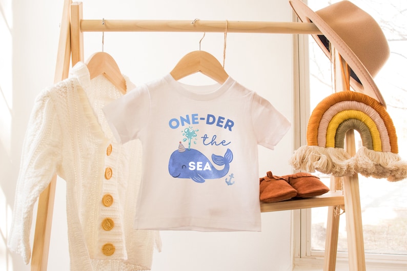 May include: White baby onesie with a blue whale wearing a party hat and the text "ONE-DER the SEA" in blue lettering.