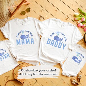 May include: A set of four white t-shirts with a blue whale graphic and the text "One-der the Sea" with a different word for each shirt: "Mama", "Daddy", "Sister", and "the Sea".