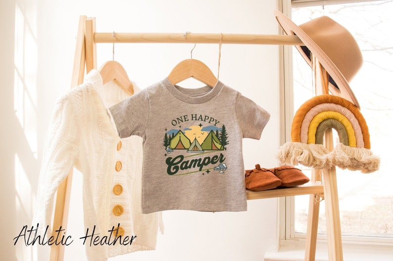 Puede incluir: Una camiseta gris de manga corta con el texto "One Happy Camper" en una fuente retro. El dise&ntilde;o presenta una escena de monta&ntilde;a con tres tiendas de campa&ntilde;a y una puesta de sol. La camiseta est&aacute; colgada en un estante de madera.