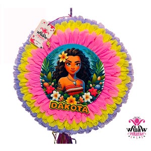 Puede incluir: Una piñata redonda con un diseño colorido. El centro presenta una ilustración de dibujos animados de una mujer con el nombre "Dakota" debajo. La piñata tiene un diseño en capas con bordes con flecos rosas, amarillos y morados. Una etiqueta con la marca "Wuaw Piñatas" está adjunta.