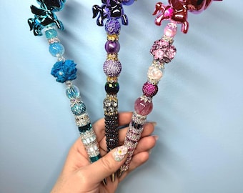 May include: Three decorative pens featuring unicorn toppers in metallic blue, dark purple, and metallic pink. Each pen is embellished with a variety of colorful beads, including textured floral shapes, sparkling rhinestones, and smooth spheres.