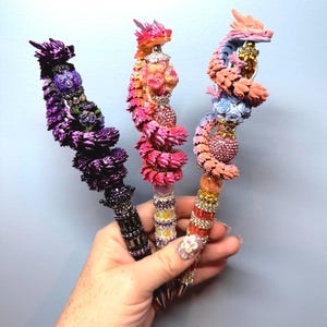 Könnte beinhalten: Drei dekorative Stifte mit skulptierten Drachenfiguren, die sich um den oberen Schaft winden. Die Stifte sind mit komplexen Texturen, funkelnden Strasssteinen und floralen Akzenten in leuchtenden Violett-, Rosa- und Pfirsichtönen verziert.