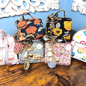 May include: A selection of miniature handbag keyrings in various floral and patterned designs. The keyrings feature gold-tone hardware, decorative charms, and a small backpack design. The colour palette includes pink, blue, black, and white.