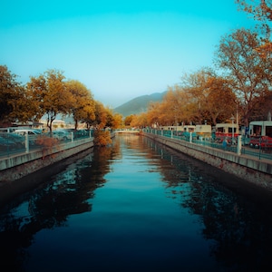 Puede incluir: Una vista panorámica de un canal con agua azul clara que refleja el cielo. Los árboles con follaje otoñal bordean las orillas, y se ven pequeños edificios y vehículos a lo largo del camino. El cielo es de un azul vibrante.
