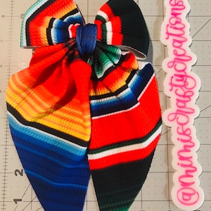 May include: A large hair bow featuring a vibrant serape pattern in red, orange, yellow, blue, and green stripes. The bow is tied with a blue center knot and has long, pointed tails. The text "@mimiscraftycreations" is visible on a white label.
