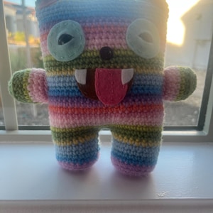 May include: A handmade crochet creature with a rectangular body, small arms, and legs. The toy features a rainbow striped pattern, large green eyes, and a felt mouth with a pink tongue. The toy is sitting on a white windowsill.