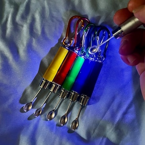 May include: Five colourful keychain tools with a silver spoon-like tip. The tools are yellow, red, green, blue, and purple. The tools are arranged in a fan shape with the tips pointing towards the bottom of the image.