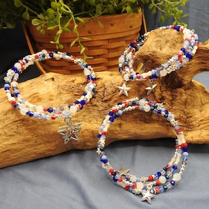 May include: Three patriotic-themed bracelets with red, white, and blue beads. Each bracelet features a silver star charm. The bracelets are displayed on a piece of driftwood, with a basket and greenery in the background.