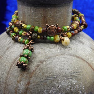 May include: A multi-strand bracelet with a cat charm. The bracelet features a mix of green, yellow, brown, and copper-colored beads. A copper cat charm and a dangling cream-colored bead add detail. The bracelet is wrapped around a gray, textured object.