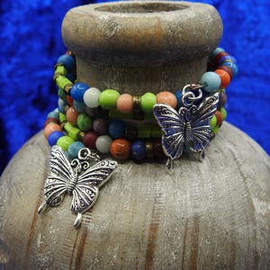 May include: Stacked beaded bracelets in various colors, including blue, green, orange, and pink, are adorned with silver butterfly charms. The bracelets are displayed on a textured, light brown surface against a vibrant blue background.