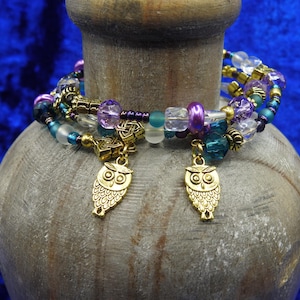 May include: A multi-strand bracelet with gold, purple, teal, and clear beads. Two gold owl charms dangle from the bracelet. The bracelet is displayed on a textured, gray surface against a deep blue background.