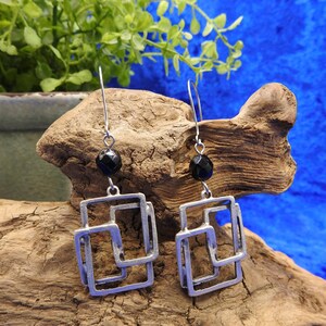May include: A pair of silver-toned earrings with a geometric square design. Each earring features a black, round bead at the top and a series of overlapping squares. The earrings are displayed against a natural wood background with a blue backdrop.