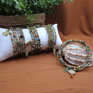 May include: Several beaded wrap bracelets in shades of blue, brown, and gold are displayed on a white pillow and a seashell. The bracelets have decorative charms, including a feather and a rose. The background is a brown fabric.