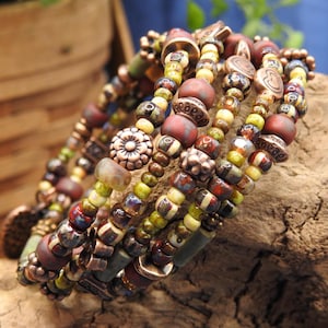 May include: A stack of beaded bracelets with a bohemian style. The bracelets feature a mix of colors, including red, green, yellow, and brown. The beads are made of various materials and shapes, including round, cylindrical, and floral designs.