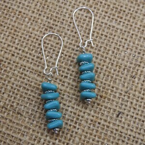 May include: A pair of turquoise blue earrings with silver-colored hooks. Each earring features a vertical stack of turquoise-colored beads, separated by small silver-colored beads. The earrings are displayed against a textured, tan background.