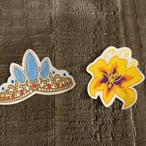 K&ouml;nnte beinhalten: Zwei Aufkleber, einer mit einer goldenen Krone mit blauen Edelsteinen und der andere mit einer gelben Blume mit einem violetten Zentrum.