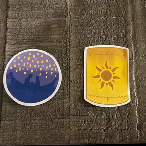 K&ouml;nnte beinhalten: Zwei runde Aufkleber, einer lila mit einer Silhouette eines Paares in einem Boot mit gelben Laternen, der andere gelb mit einem Sonnendesign.