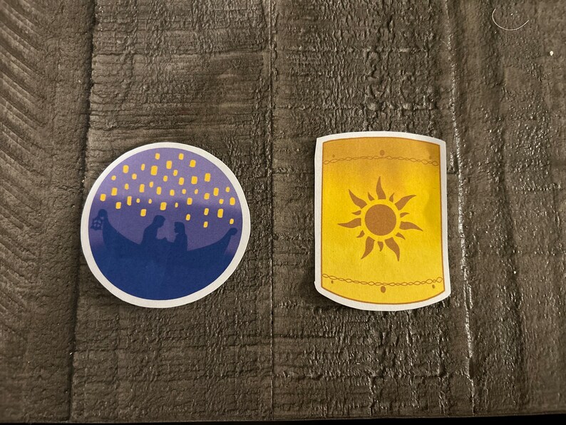 K&ouml;nnte beinhalten: Zwei runde Aufkleber, einer lila mit einer Silhouette eines Paares in einem Boot mit gelben Laternen, der andere gelb mit einem Sonnendesign.