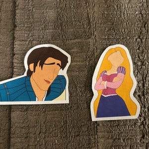 K&ouml;nnte beinhalten: Zwei Comic-Aufkleber eines Prinzen und einer Prinzessin aus dem Disney-Film Rapunzel - Neu verf&ouml;hnt. Der Prinz tr&auml;gt ein blaues und goldenes Outfit und hat braunes Haar. Die Prinzessin hat lange blonde Haare und tr&auml;gt ein lila Kleid mit einem rosafarbenen Oberteil.