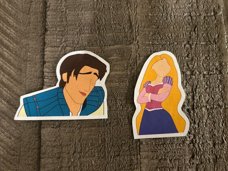 K&ouml;nnte beinhalten: Zwei Comic-Aufkleber eines Prinzen und einer Prinzessin aus dem Disney-Film Rapunzel - Neu verf&ouml;hnt. Der Prinz tr&auml;gt ein blaues und goldenes Outfit und hat braunes Haar. Die Prinzessin hat lange blonde Haare und tr&auml;gt ein lila Kleid mit einem rosafarbenen Oberteil.