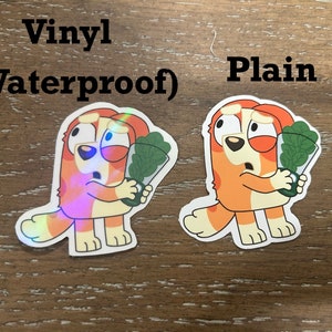 K&ouml;nnte beinhalten: Zwei Cartoon-Hunde-Aufkleber, einer mit holografischem Finish und der andere mit einfachem Finish. Beide Aufkleber zeigen einen Hund, der einen Strau&szlig; gr&uuml;ner Bl&auml;tter h&auml;lt. Der Text "Vinyl (Waterproof)" befindet sich &uuml;ber dem holografischen Aufkleber und der Text "Plain" &uuml;ber dem einfachen Aufkleber.