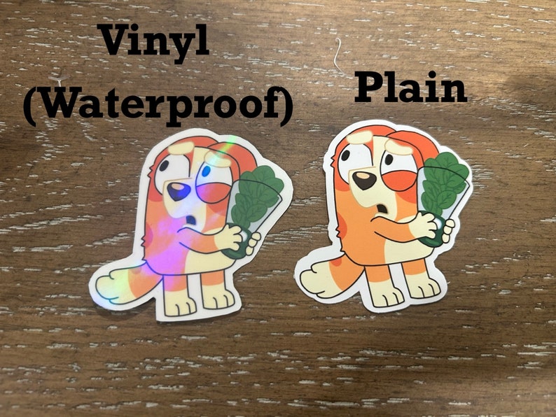 K&ouml;nnte beinhalten: Zwei Cartoon-Hunde-Aufkleber, einer mit holografischem Finish und der andere mit einfachem Finish. Beide Aufkleber zeigen einen Hund, der einen Strau&szlig; gr&uuml;ner Bl&auml;tter h&auml;lt. Der Text "Vinyl (Waterproof)" befindet sich &uuml;ber dem holografischen Aufkleber und der Text "Plain" &uuml;ber dem einfachen Aufkleber.