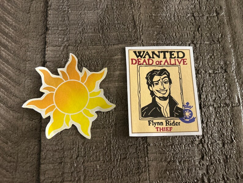 K&ouml;nnte beinhalten: Ein Fahndungsplakat mit einer Cartoon-Illustration von Flynn Rider, einem Dieb, mit dem Text "WANTED DEAD or ALIVE" und "Flynn Rider THIEF". Das Plakat befindet sich neben einer gelben Sonne mit Strahlen.