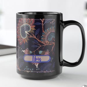 Può includere: Tazza in ceramica nera con un motivo floreale nei toni del marrone, blu e verde. La tazza presenta la parola "Ibu" in un rettangolo azzurro. La tazza ha un manico grande ed è appoggiata su una superficie bianca.