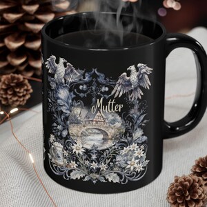 May include: Black ceramic mug with a decorative design featuring eagles, a bridge, and floral elements. The word "Mutter" is printed on the mug. The mug is filled with a hot beverage, and steam is rising from it.