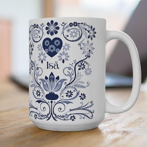 Peut inclure: Mug en céramique blanche avec un motif floral bleu et le mot "Isä". Le motif comprend un cœur et divers motifs floraux. Le mug est posé sur une surface en bois, avec un ordinateur portable en arrière-plan.