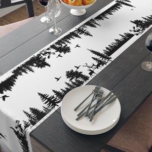 Puede incluir: Un camino de mesa blanco y negro con un diseño de silueta de bosque. El camino de mesa está sobre una mesa de madera oscura con un tazón de manzanas, dos copas de vino y un plato con cubiertos.