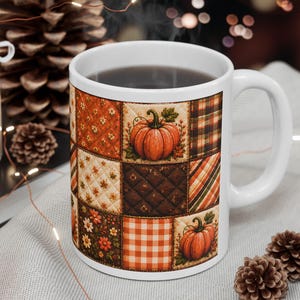 May include: A white ceramic mug featuring a patchwork quilt design with various autumn-themed patterns including orange pumpkins, brown quilted squares, plaid, and floral motifs. The mug has a standard handle and a smooth, glossy finish on the exterior.