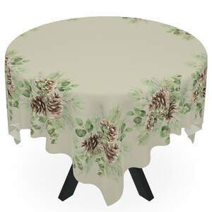 Pode incluir: Uma toalha de mesa redonda com um design de pinhas e folhas de eucalipto verdes e marrons sobre um fundo bege. A toalha de mesa está drapeada sobre uma mesa com pernas pretas.