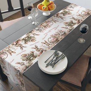 May include: White table runner with a repeating pattern of bunnies, baskets of eggs, and the text "Happy Easter". The runner is on a dark wood table with a bowl of apples and two glasses of wine.