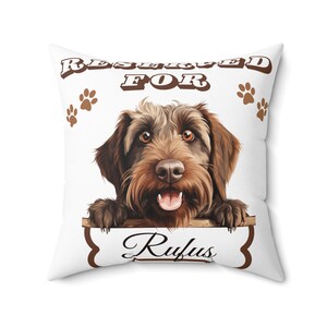 May include: Square white cushion featuring a brown dog illustration with the text "RESERVED FOR Rufus". The dog has brown fur and is smiling. Brown paw prints are scattered around the text.