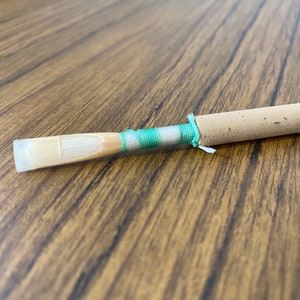 May include: A brown and white clarinet reed with a green and white string tied around it.