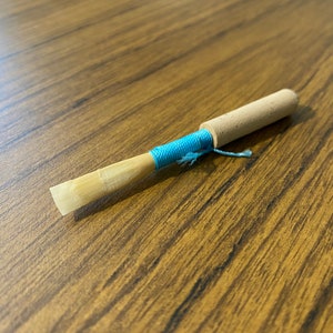 May include: A light brown wooden clarinet reed with a blue string tied around it.