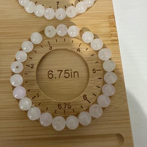May include: Two rose quartz beaded bracelets, each with round beads, are displayed on a wooden measuring board. One bracelet is 7 inches in diameter, and the other is 6.75 inches. The beads are a soft pink color.