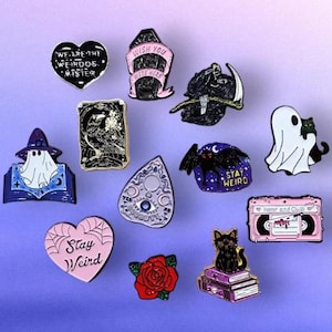 Pode incluir: Uma coleção de alfinetes de esmalte com um tema assustador. Os designs incluem um fantasma, um ceifador, um gato preto, uma tábua Ouija, uma fita cassete e alfinetes em forma de coração com texto como "Stay Weird". Os alfinetes são de várias cores.