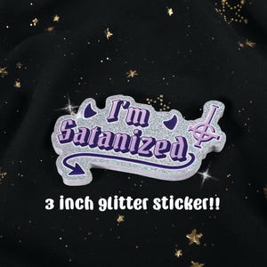 May include: A 3-inch glitter sticker with the text "I'm Satanized" in purple and silver. The sticker has a unique shape with a stylized symbol and crescent moon accents. The background is black with gold glitter.